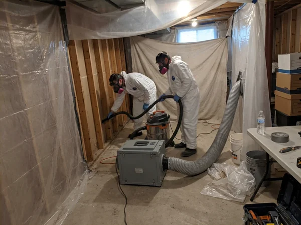 HEPA air scrubber running while two technicians HEPA-vacuum exposed studs