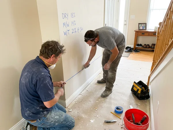 Two technicians making a measured flood cut along drywall marked with moisture readings